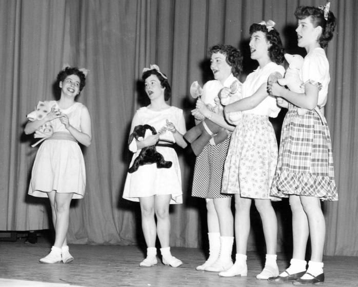 Picture of female students on stage dressed as little girls and holding stuffed animals -- singing