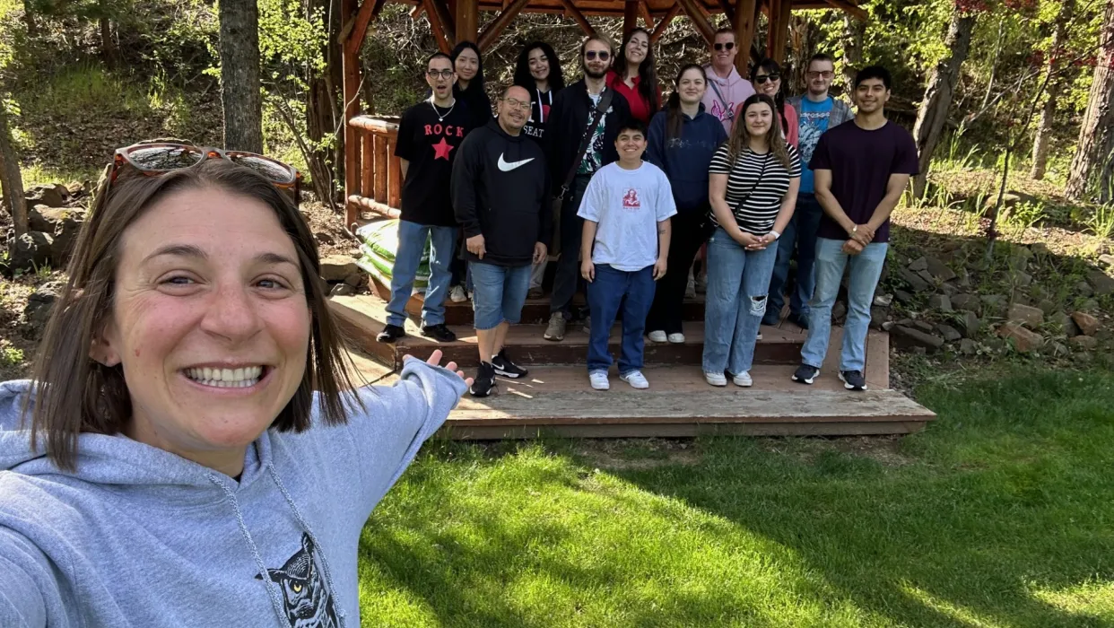 Person with OIT sweatshirt takes selfie with group of OIT students in background. Outdoor space with green grass. 